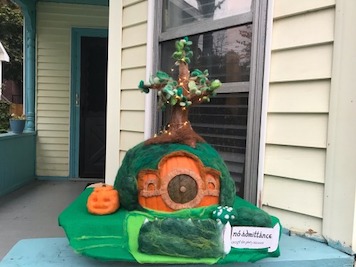 A pumpkin covered in felt and felting wool to depict Bag End. The stem has been turned into a tree that is lit up with lights. There is a round door colored on the front and a sign that reads 'No Admittance Except on Party Business' in the front next to a mossy fence. The garden has small green shrubs and a bush covered in white flowers. There is also a small needle felted jack o lantern in the garden. The pumpkin is outside on Rebecca's porch.