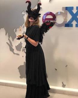 Rebecca dressed as a winged demon in all black with red tipped horns standing in front of the edX logo in the edX Office October 2019. She is holding a small skeleton trophy. The wings have red feathers at the top.